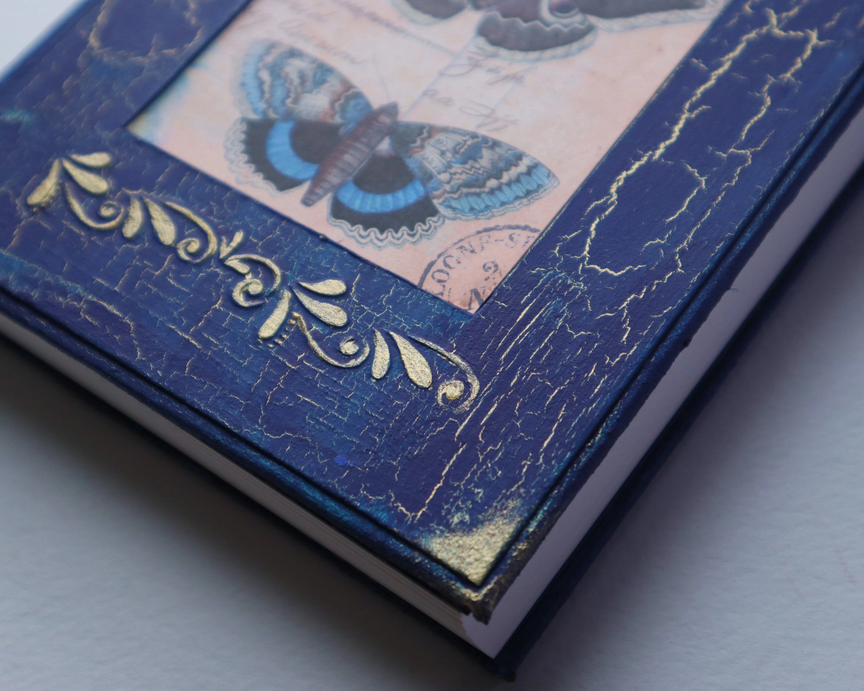 The cover of a journal. It is royal blue, the paint is cracking and gold shows through the cracks. There is a photo of 3 blue butterflies in the center. Under the photo there is a gold embossed elegant pattern, as well as gold in all 4 corners.