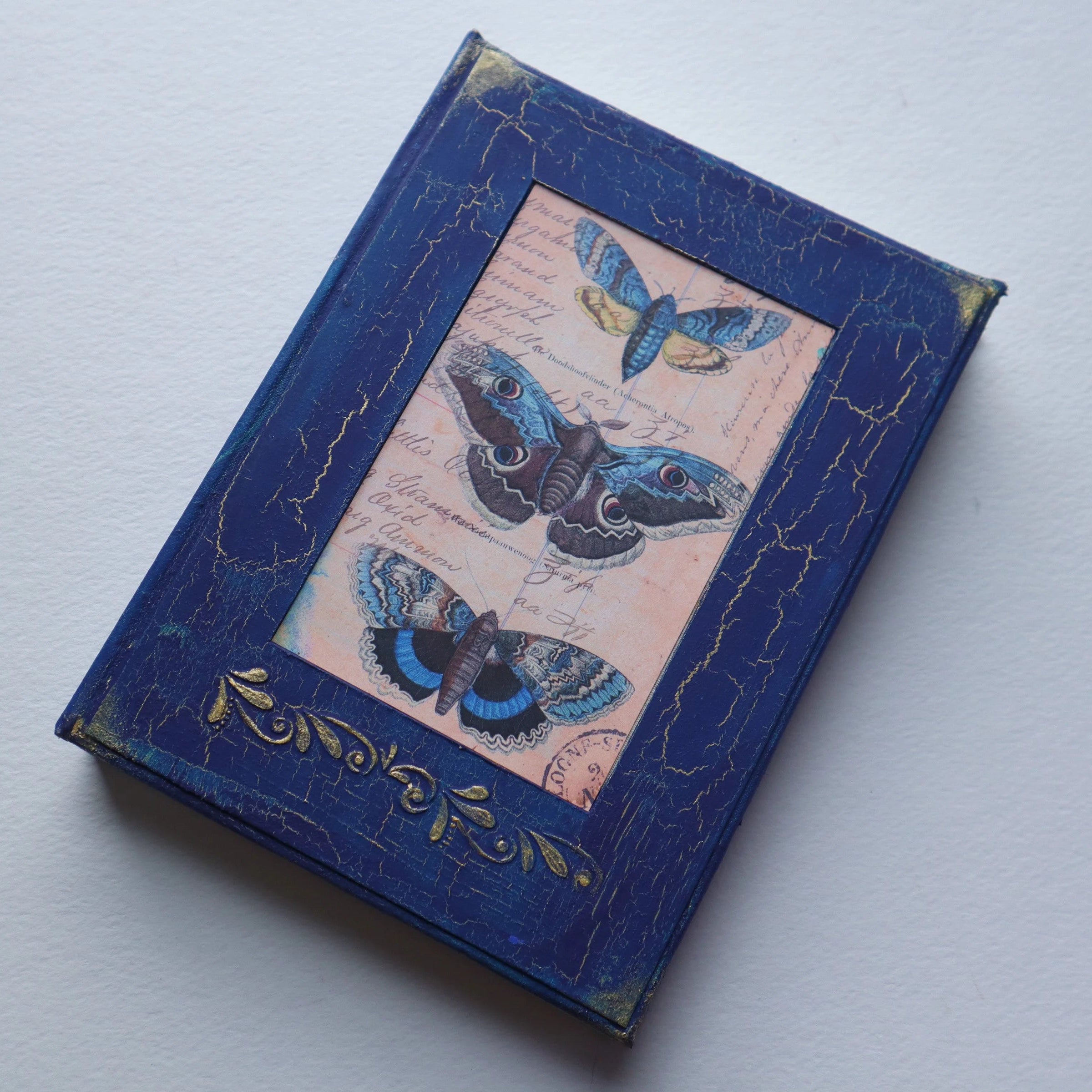The cover of a journal. It is royal blue, the paint is cracking and gold shows through the cracks. There is a photo of 3 blue butterflies in the center. Under the photo there is a gold embossed elegant pattern, as well as gold in all 4 corners.