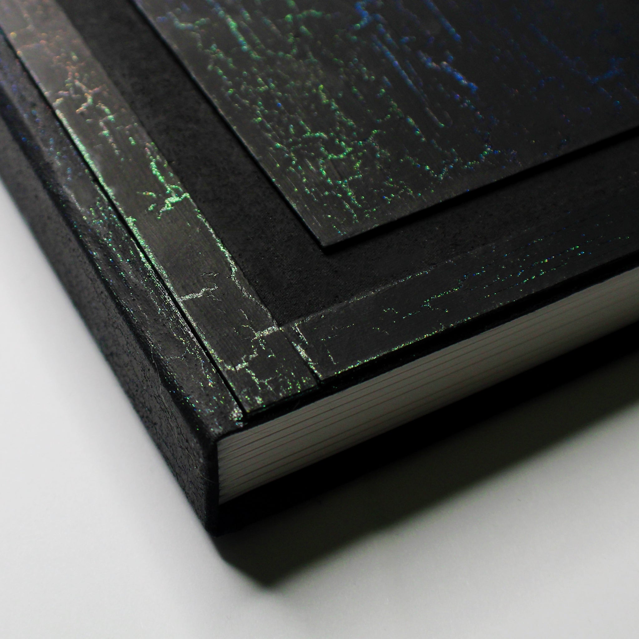The bottom left corner of a black journal with a colorful, crackled pattern on the cover. The cover has a 1/2 inch border and a 1/2 inch recessed area that surrounds a rectangular flat central panel.