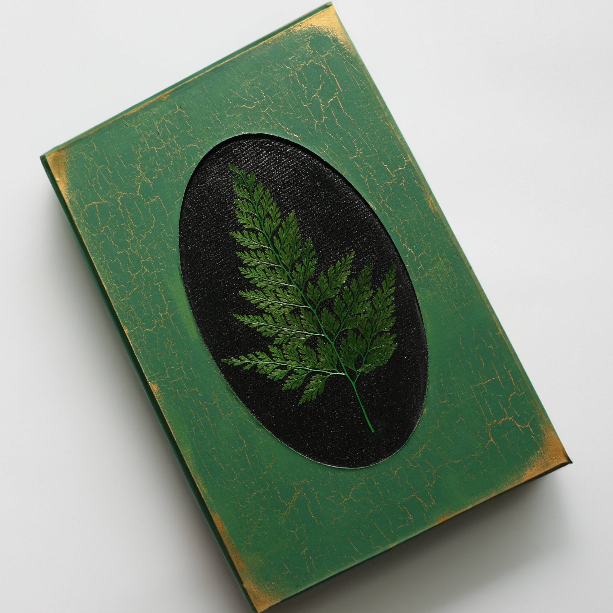A green journal with gold crackle and gold corners, with a pressed dried fern inside of a black recessed oval frame in the center.
