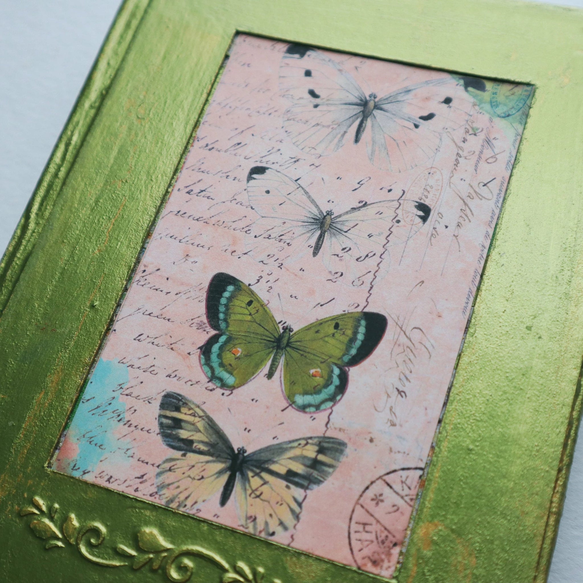 The cover of a journal. It is metallic green, the paint is cracking and gold shows through the cracks. There is a photo of 4 butterflies in the center, 2 white, 1 green, 1 yellow. Under the photo there is an embossed elegant pattern.