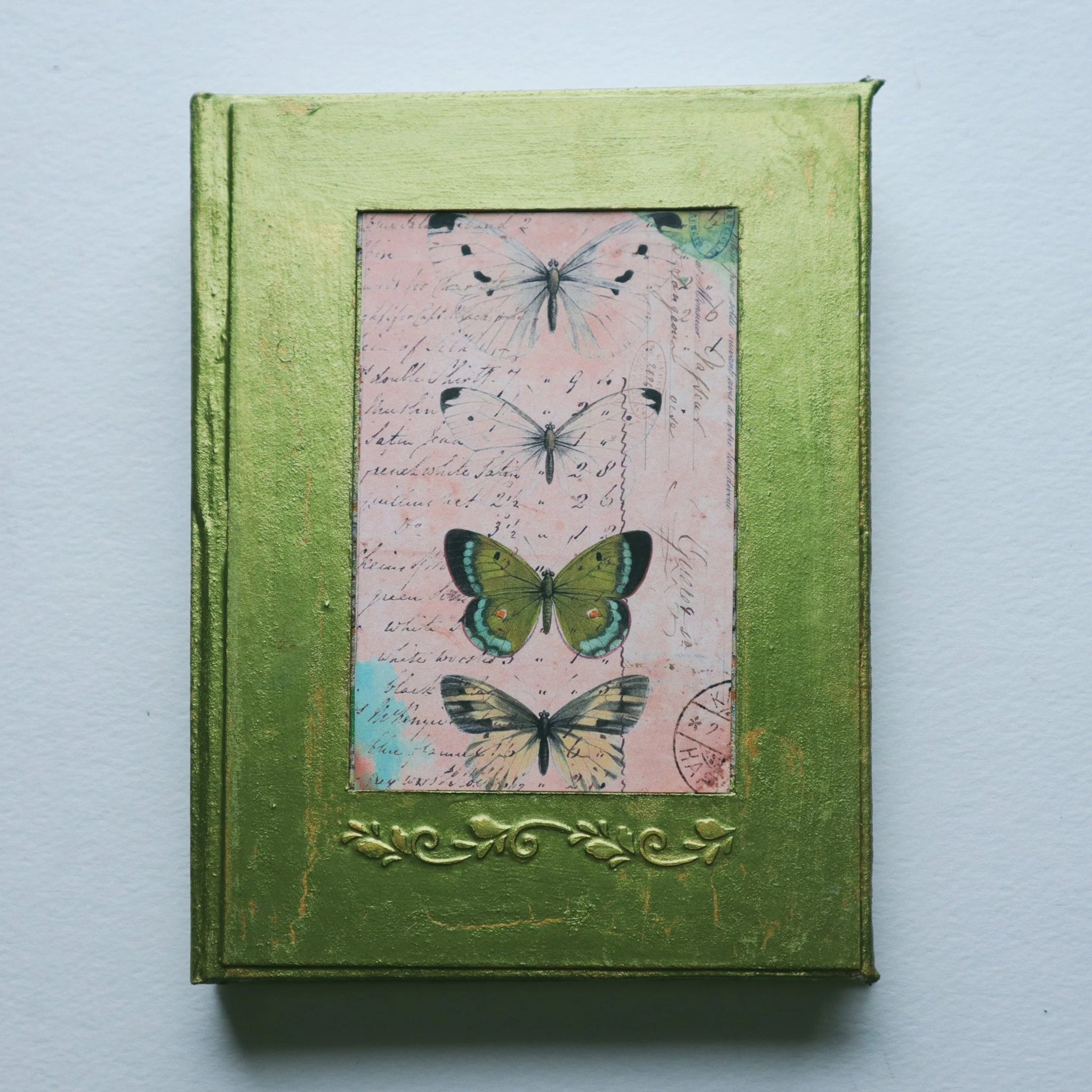 The cover of a journal. It is metallic green, the paint is cracking and gold shows through the cracks. There is a photo of 4 butterflies in the center, 2 white, 1 green, 1 yellow. Under the photo there is an embossed elegant pattern.