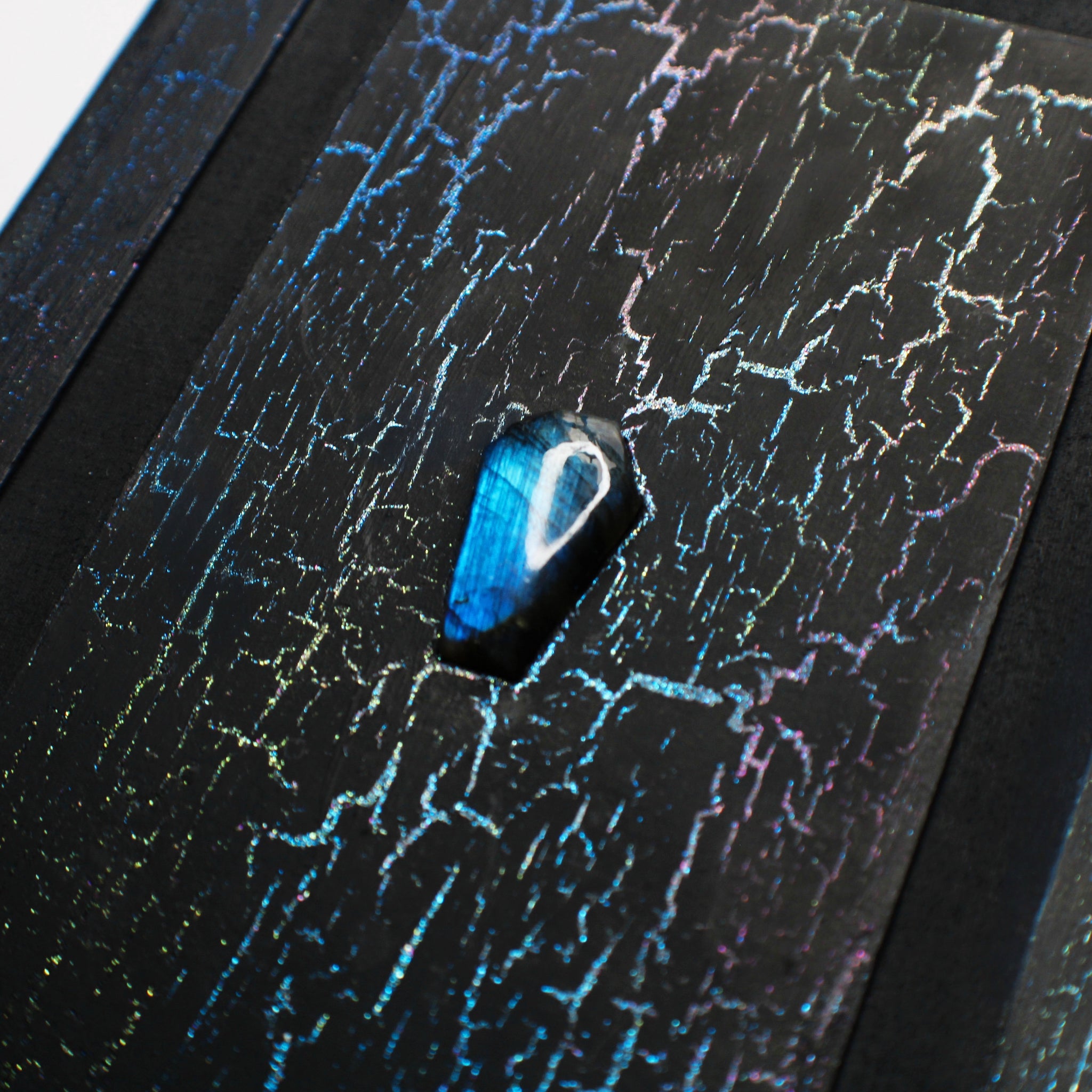 A close up image of a black journal with a colorful, crackled pattern on the cover. The cover has a 1/2 inch border and a 1/2 inch recessed area that surrounds a rectangular flat central panel, with a coffin-shaped labradorite crystal in the center that the image focuses on.