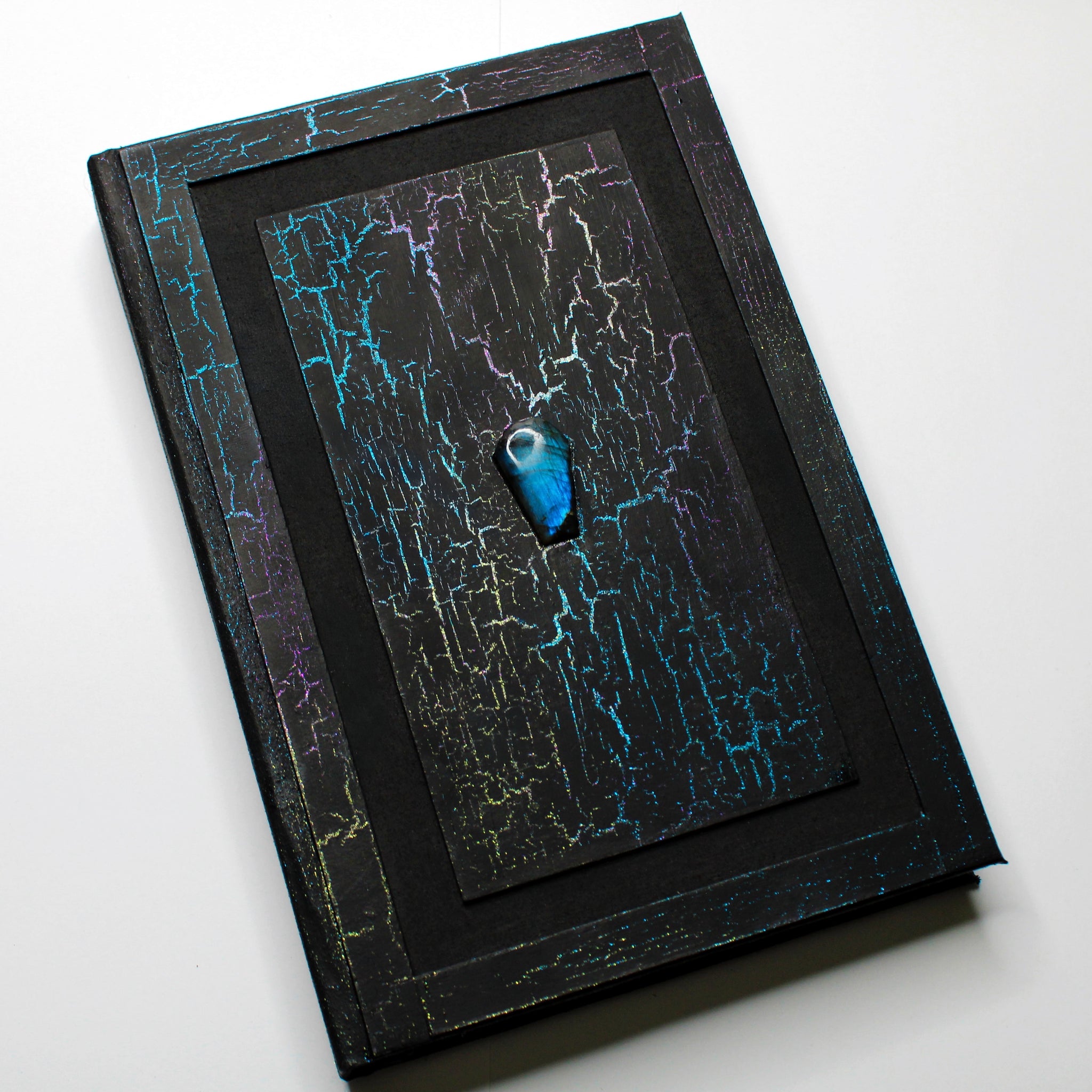 A black journal with a colorful, crackled pattern on the cover. The cover has a 1/2 inch border and a 1/2 inch recessed area that surrounds a rectangular flat central panel with a coffin-shaped labradorite crystal in the center.