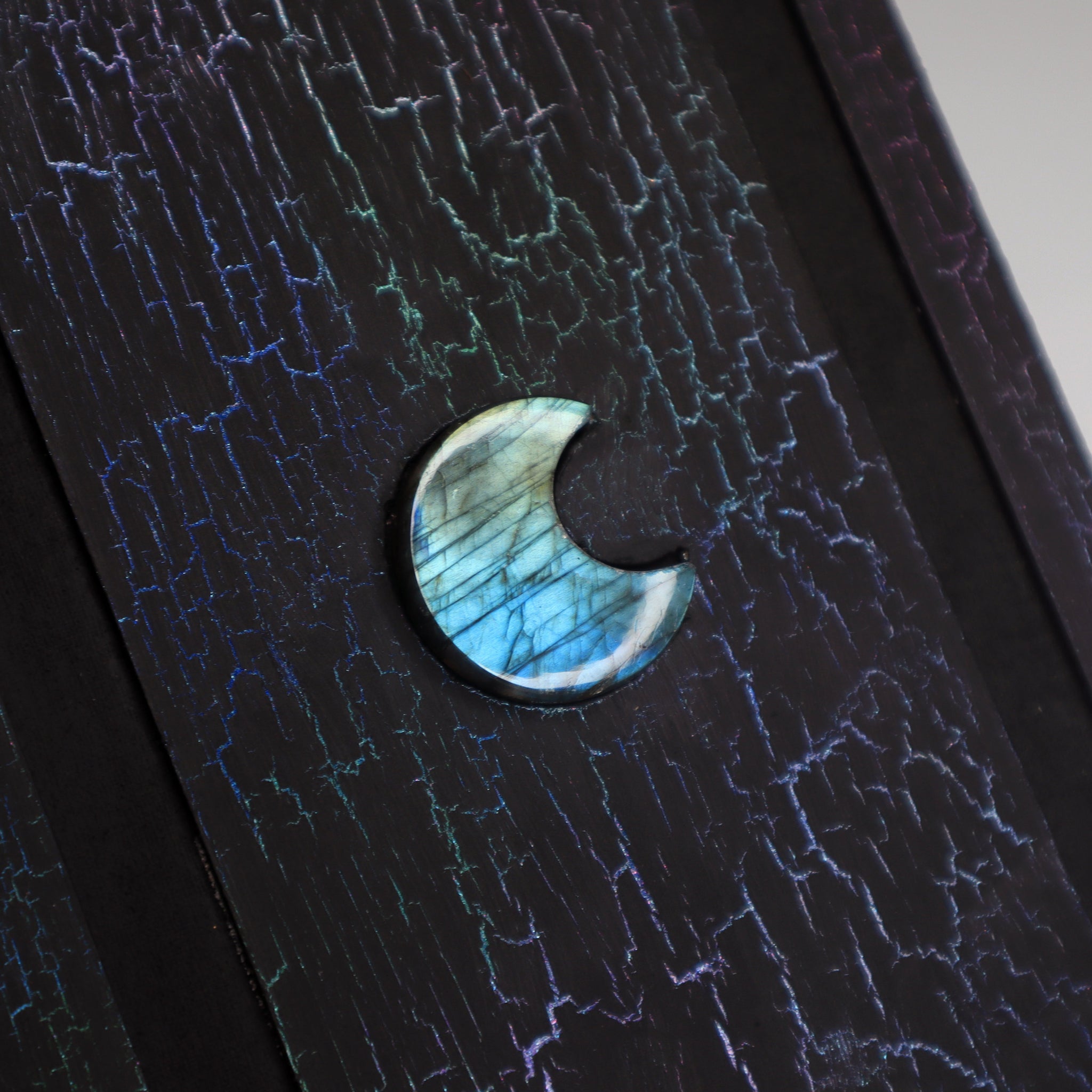 A close up of black journal with a colorful, crackled pattern on the cover. The cover has a 1/2 inch border and a 1/2 inch recessed area that surrounds a rectangular flat central panel with a coffin-shaped labradorite crystal in the center.
