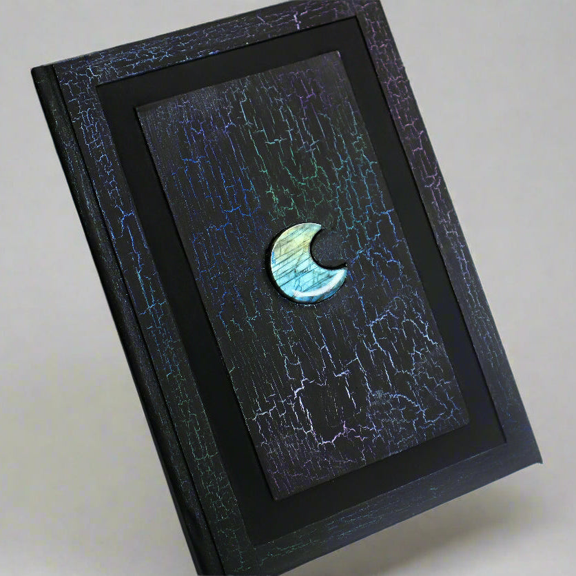 A black journal with a colorful, crackled pattern on the cover. The cover has a 1/2 inch border and a 1/2 inch recessed area that surrounds a rectangular flat central panel with a crescent moon-shaped labradorite crystal in the center.