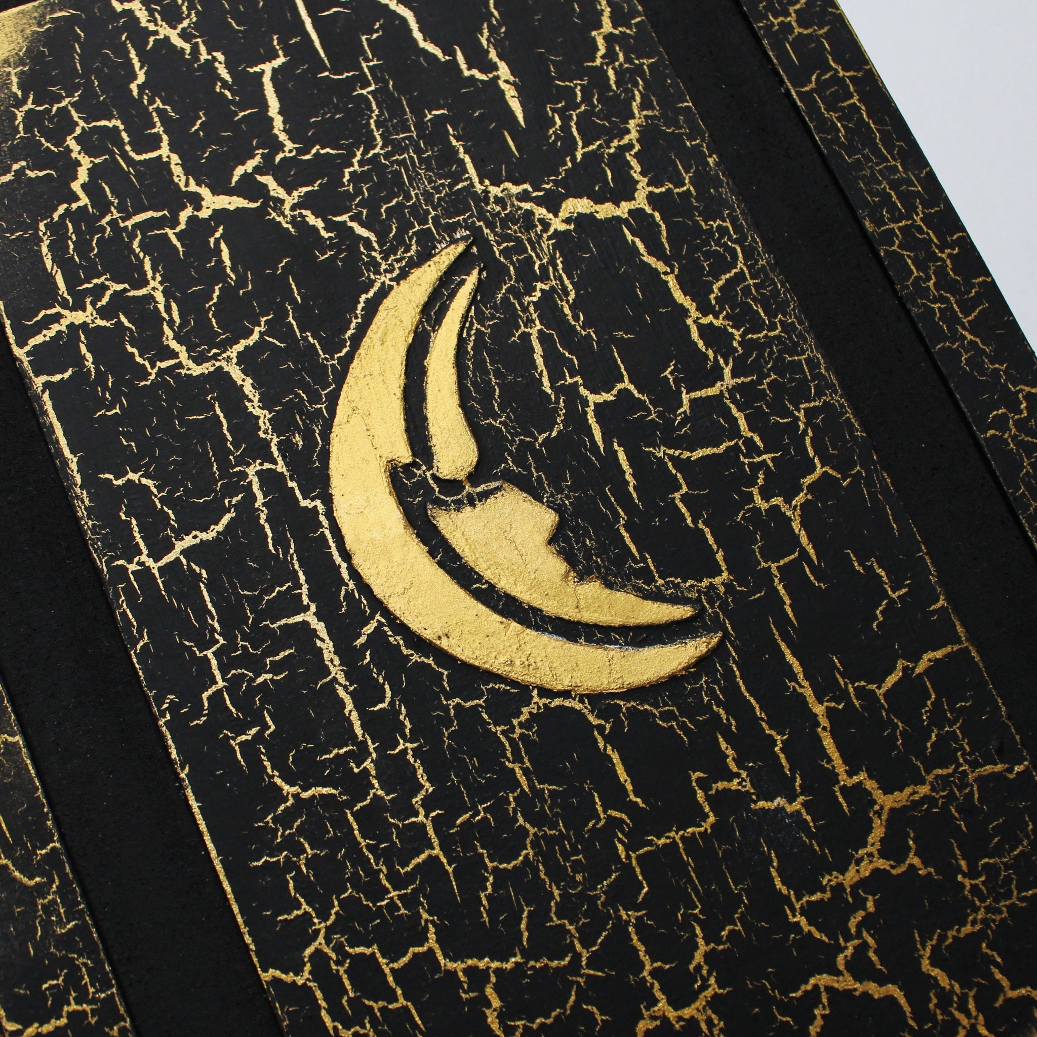 A black journal with a gold, crackled pattern on the cover. The cover has a 1/2 inch border, and a black 1/2 inch recessed area that surrounds a rectangular flat central panel with an embossed golden moon in the center which is the main focus of the image.
