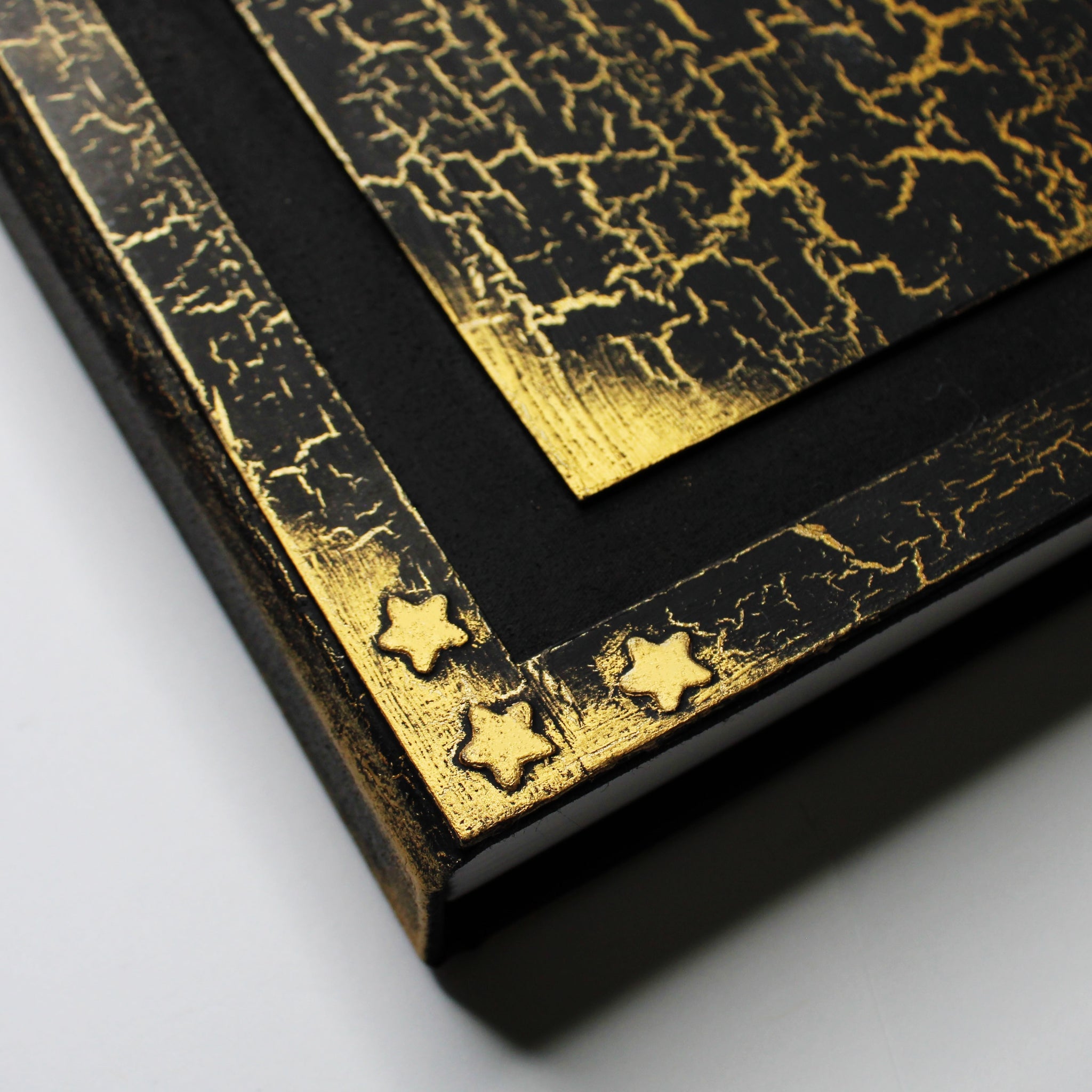 A black journal with a gold, crackled pattern on the cover. The cover has a 1/2 inch border with gold corners and 3 embossed gold stars in the corner, and a black 1/2 inch recessed area that surrounds a rectangular flat central panel with golden edges. The image focuses on the bottom left corner, showing detail in the 3 gold stars.
