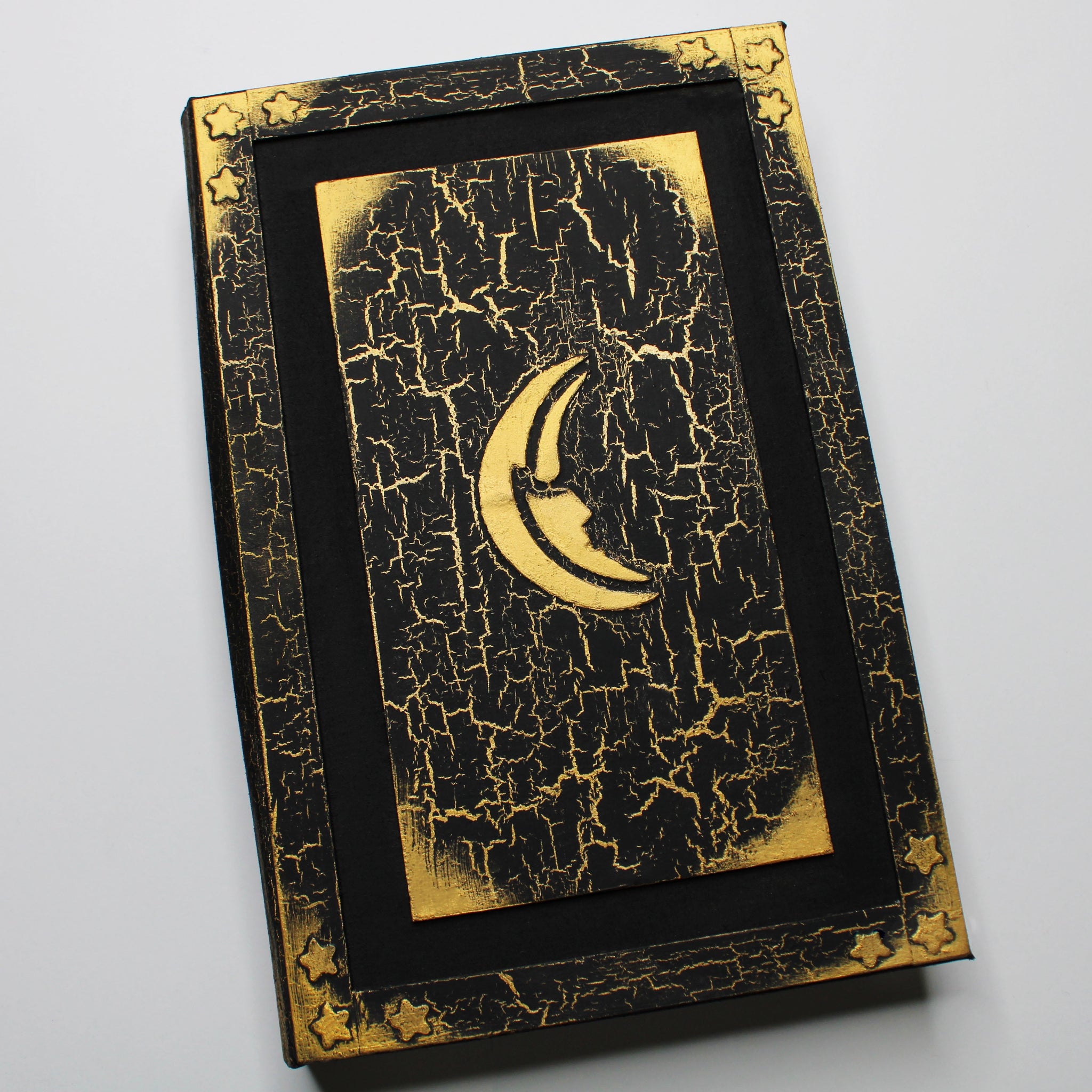 A black journal with a gold, crackled pattern on the cover. The cover has a 1/2 inch border with gold edges and 3 golden stars in each corner, and a black 1/2 inch recessed area that surrounds a rectangular flat central panel with gold corners and an embossed golden moon in the center.
