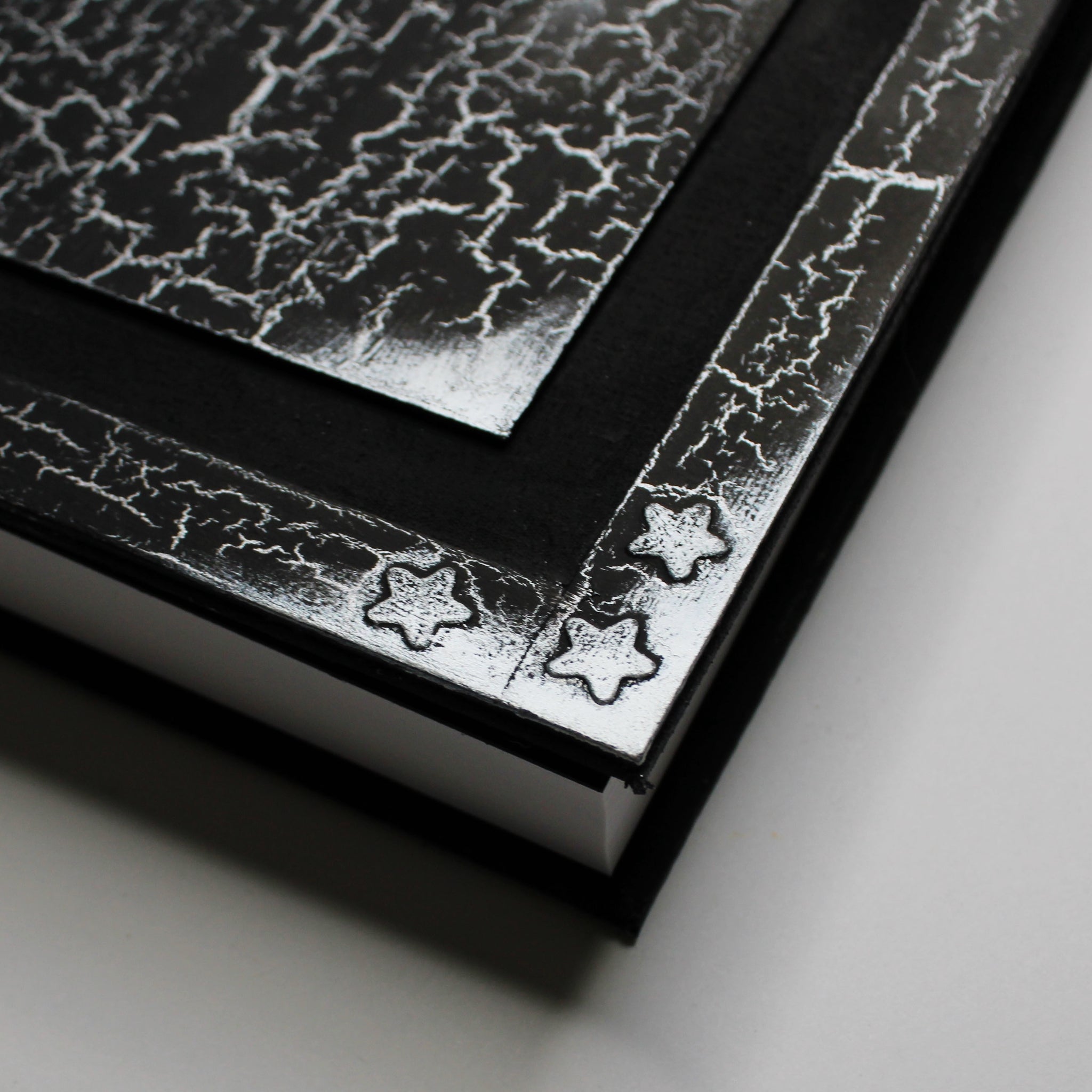 A black journal with a silver, crackled pattern on the cover. The cover has a 1/2 inch border with silver corners and 3 embossed silver stars in the corner, and a black 1/2 inch recessed area that surrounds a rectangular flat central panel with silver edges. The image focuses on the bottom left corner, showing detail in the 3 silver stars.